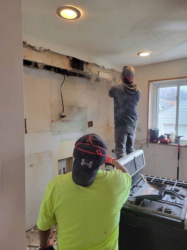 Two workers repair a damaged wall inside a room, with one standing on a ladder near a window.