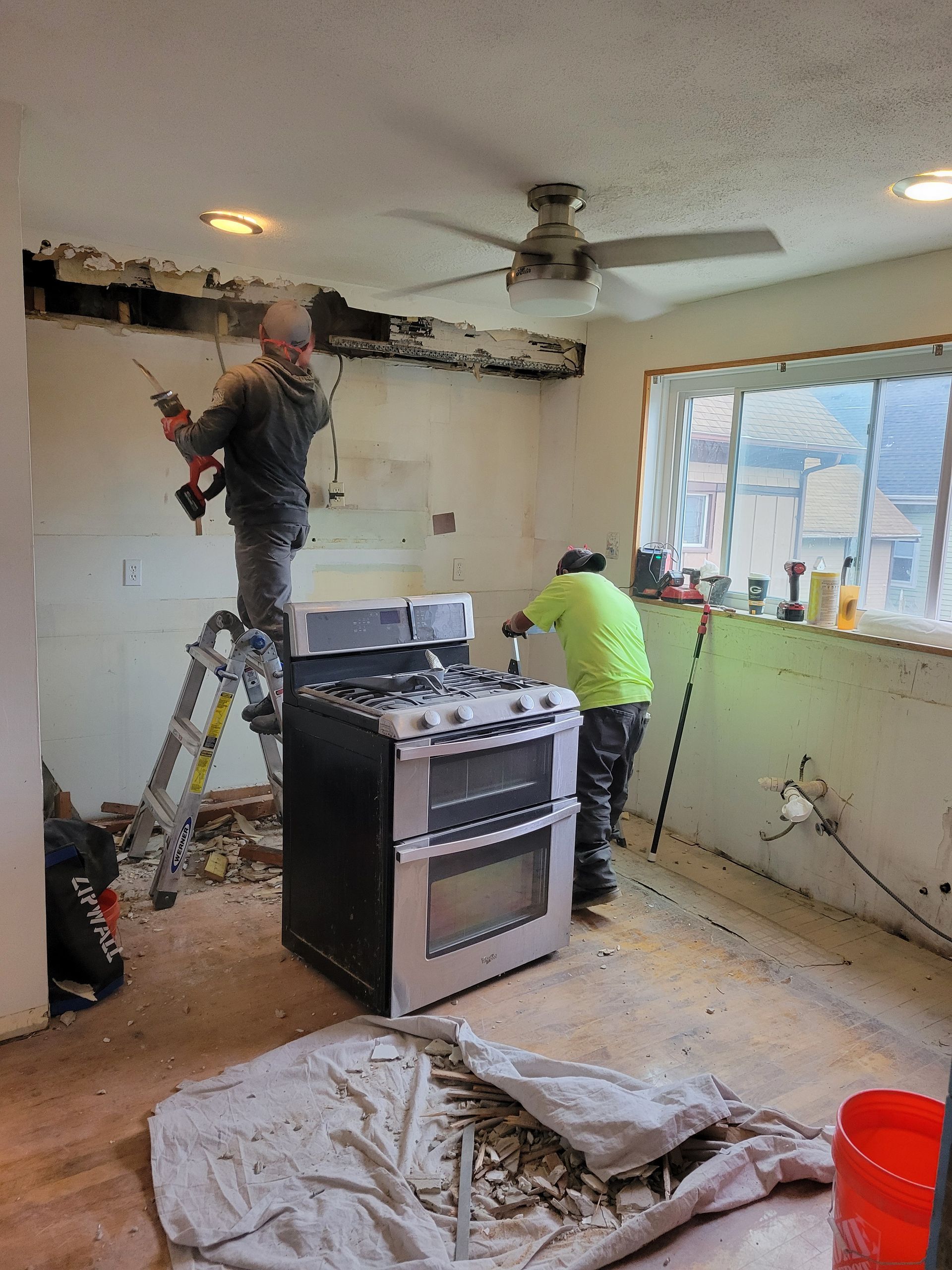 Two workers renovating a kitchen, one on a ladder and one near a stove, with debris and tools on the floor