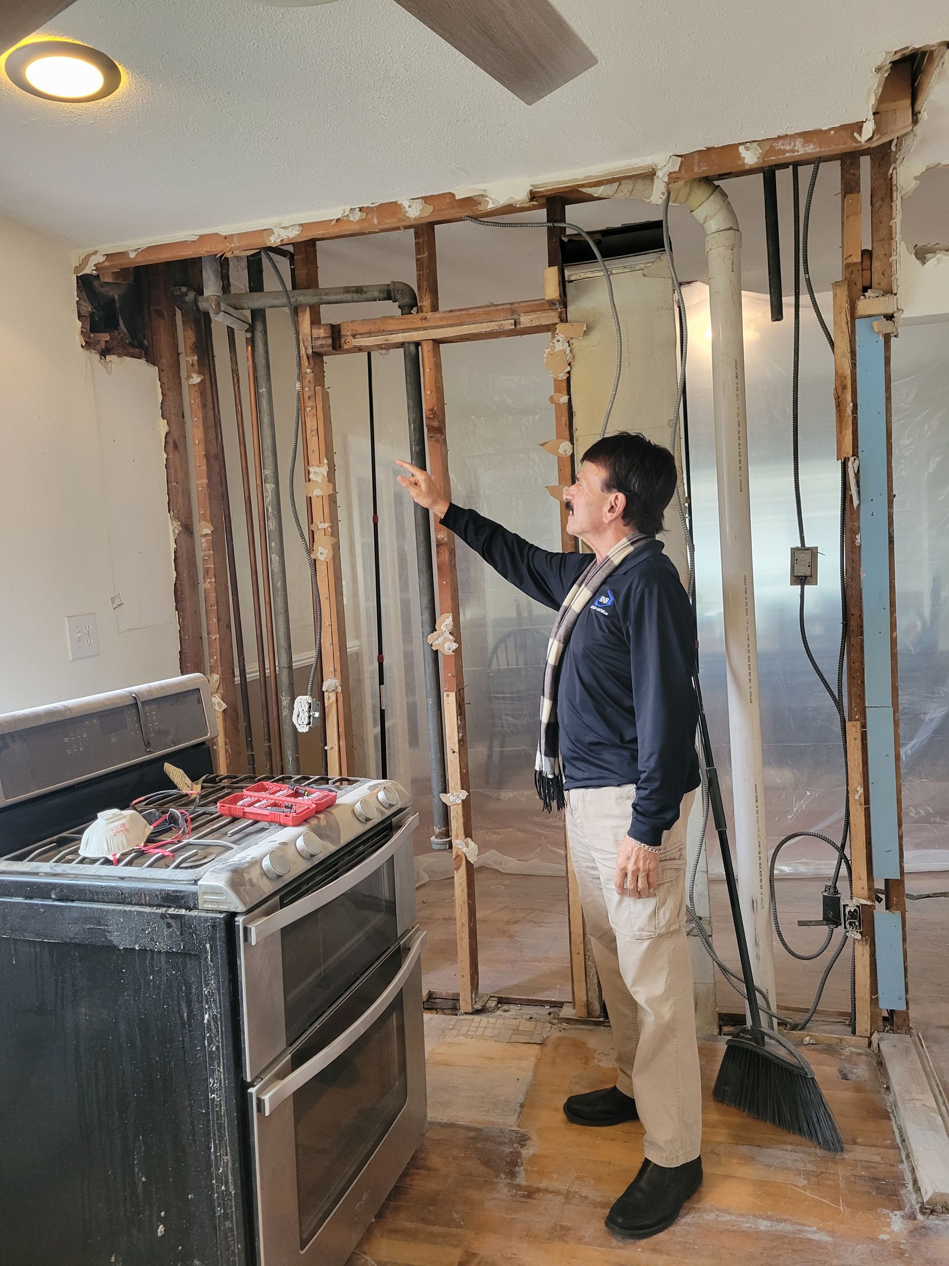 Man in a kitchen stands beside exposed wall studs and wiring during demolition.