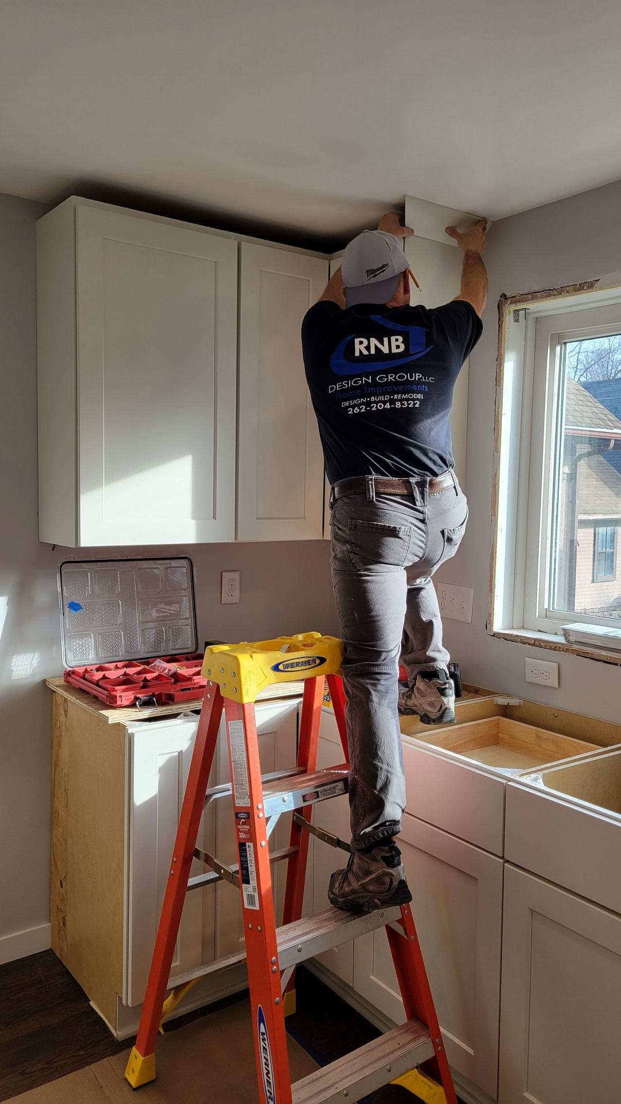Person on a red ladder installing a cabinet in a bright kitchen near a window