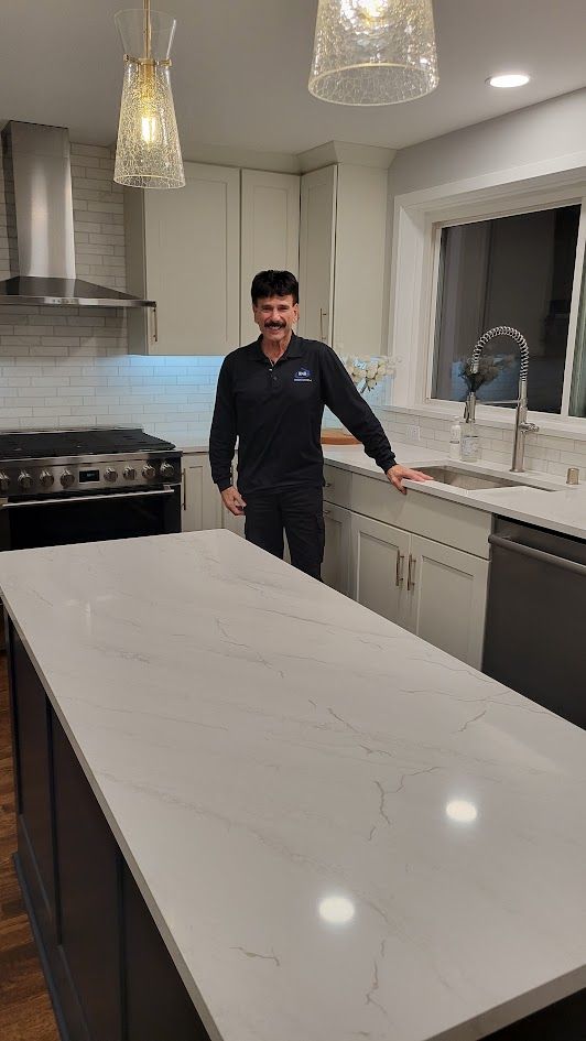 Man standing in a modern kitchen beside a white island with pendant lights and gray cabinets.