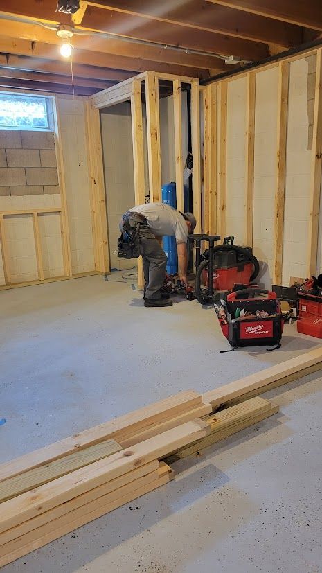 Basement under renovation with exposed studs, tools, lumber, and a worker bending near the wall
