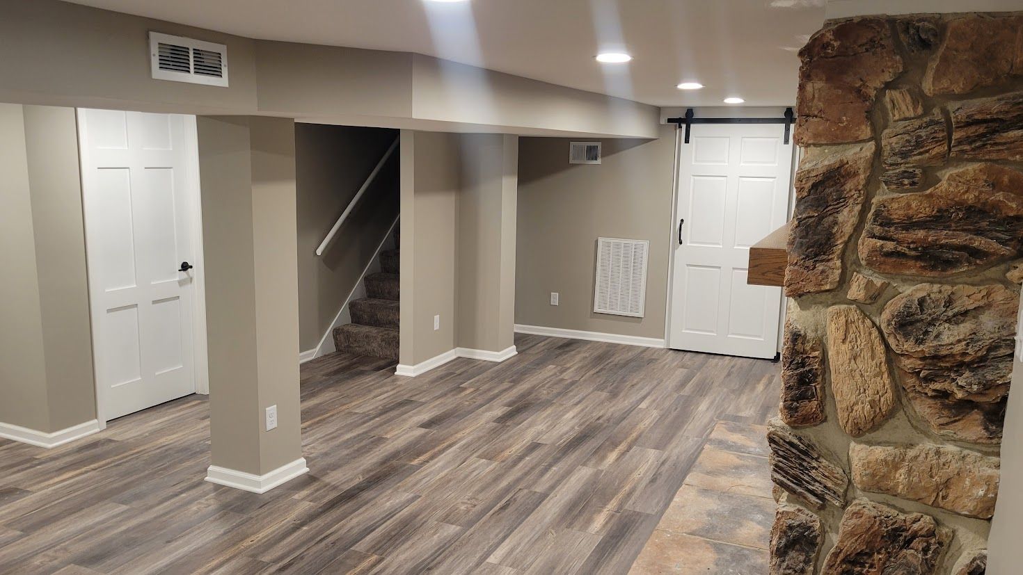 Empty basement room with wood-look flooring, white walls, staircase, and stone fireplace column
