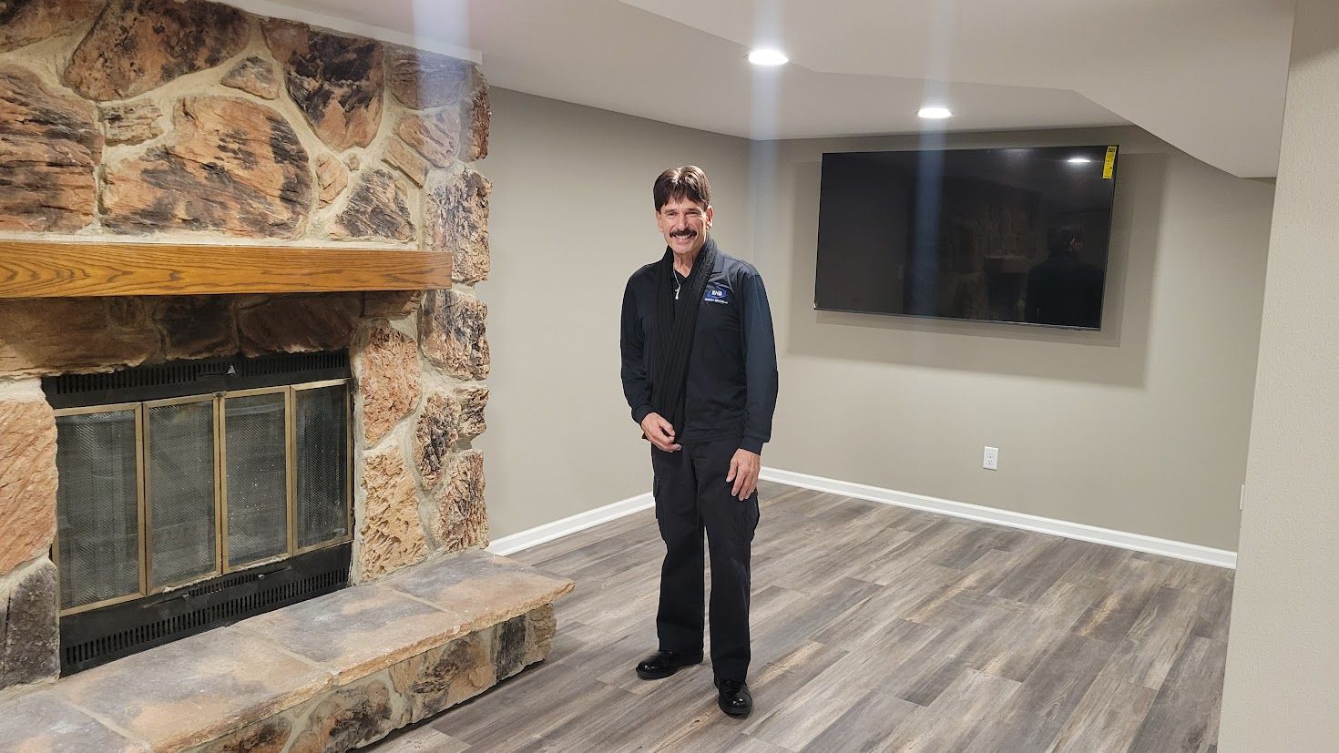 Man standing in a living room beside a stone fireplace and wall-mounted TV on gray wood flooring