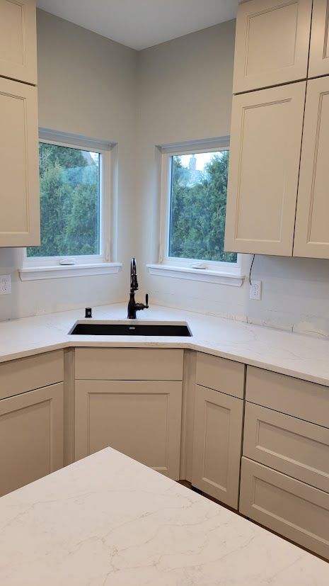 Bright modern kitchen corner with white cabinets, white countertops, and two windows above a black sink.