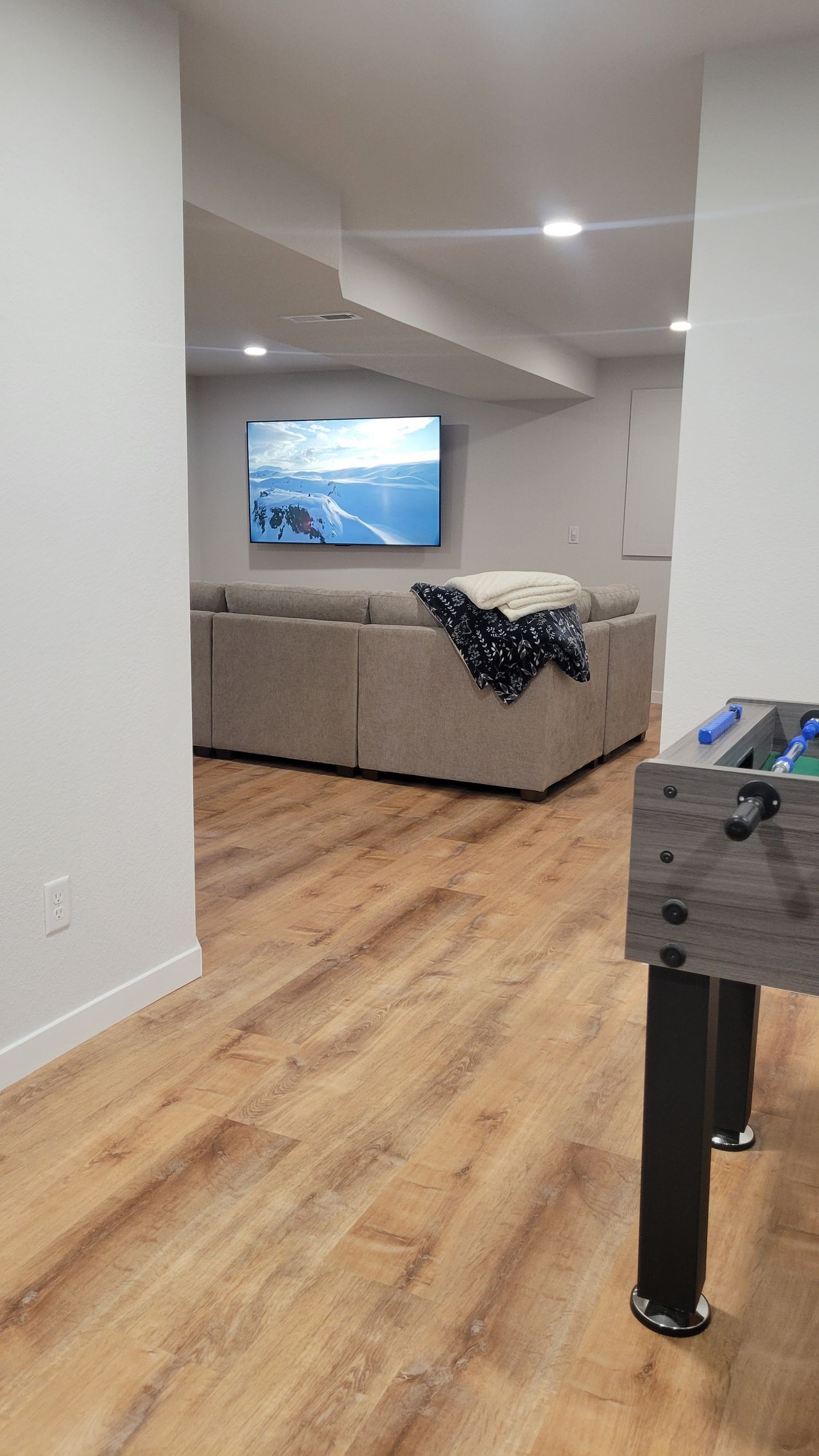 Living room with gray sectional sofa, wall-mounted TV, and a foosball table in the foreground