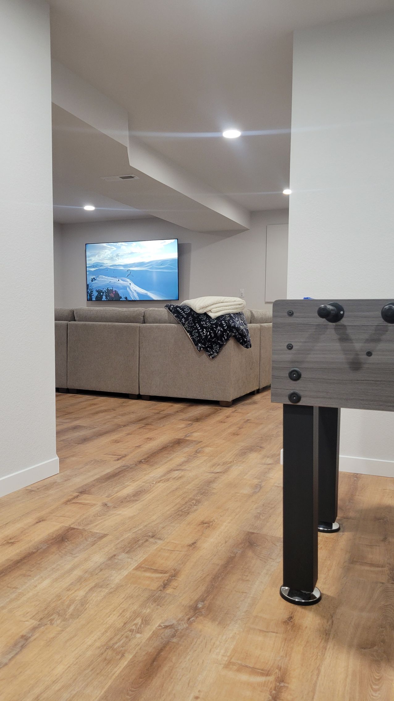 Modern basement room with wood floor, gray sectional, wall TV, and foosball table in foreground