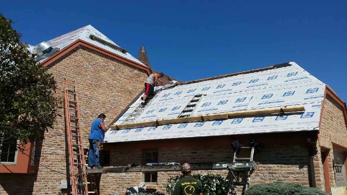 Worker installing shingles on a sloped roof beside a brick house on a sunny day