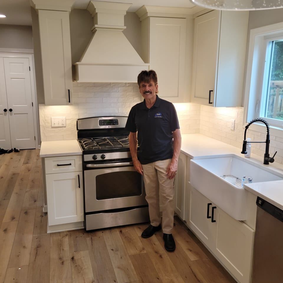 Man standing in a bright white kitchen beside a stainless steel stove and farmhouse sink.
