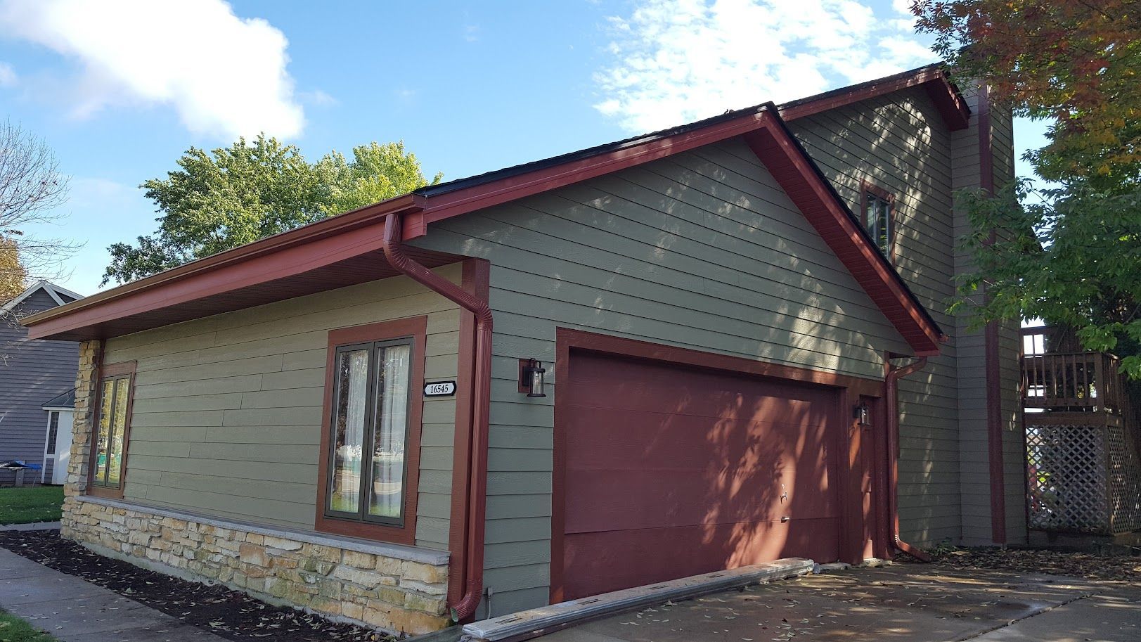 Tan house with red trim and garage door, viewed from the side on a tree-lined street.