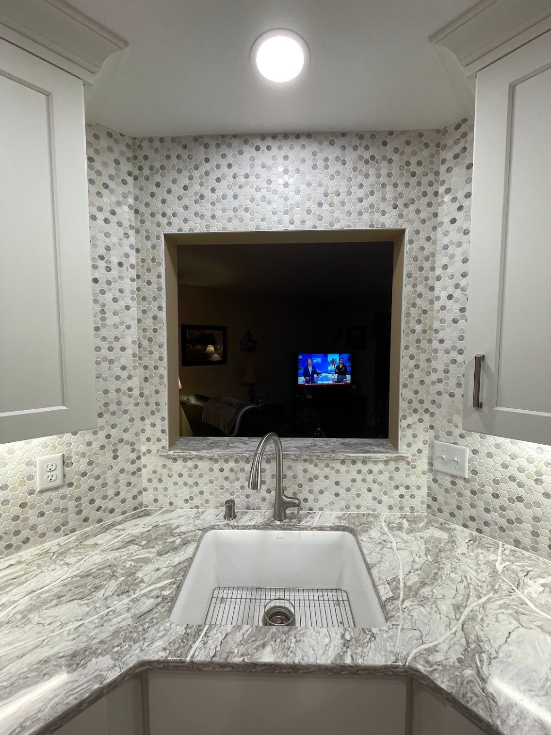 Modern bathroom with marble-patterned walls and a sink overlooking a dark room with a TV in the distance