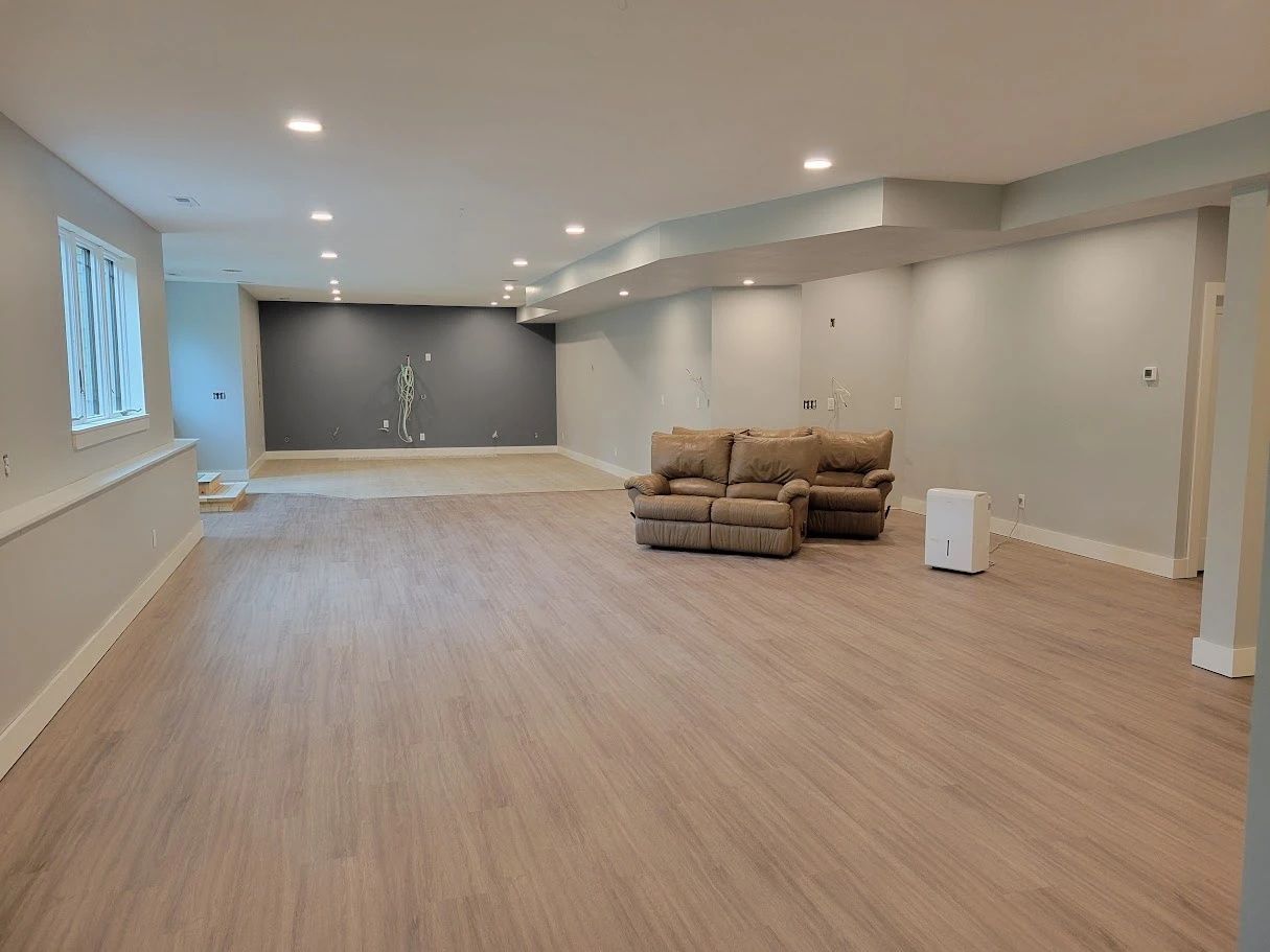Spacious empty living room with light wood floors, gray walls, recessed lights, and a beige sofa.