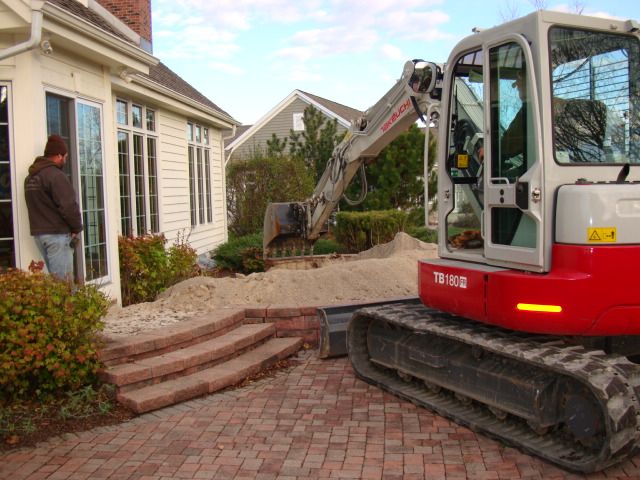 Excavator working beside a house, digging up a garden bed near brick steps.