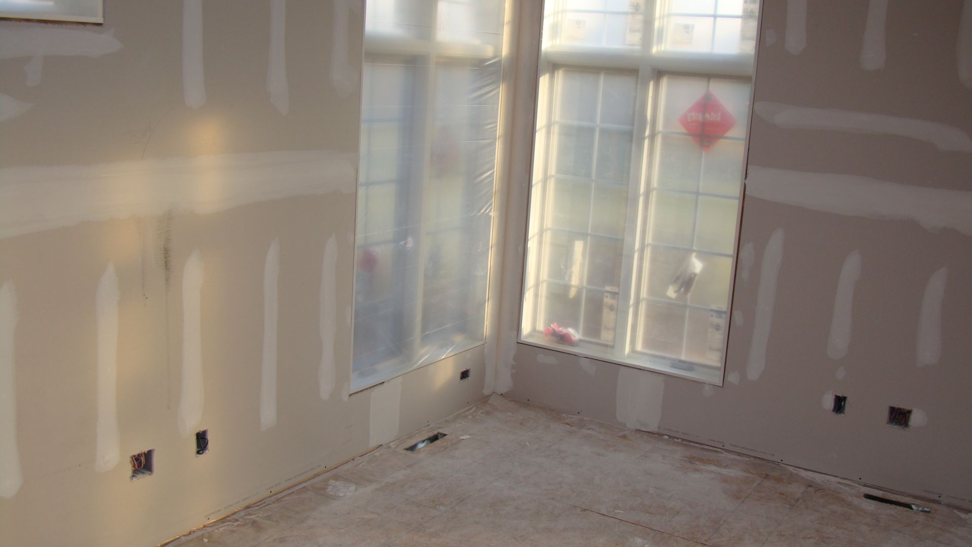 Empty room under renovation with unfinished drywall, bare floor, and sunlight through windows.