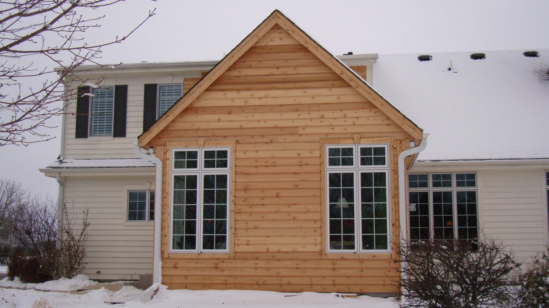 Snowy house exterior with a tan wooden sunroom extension and large windows