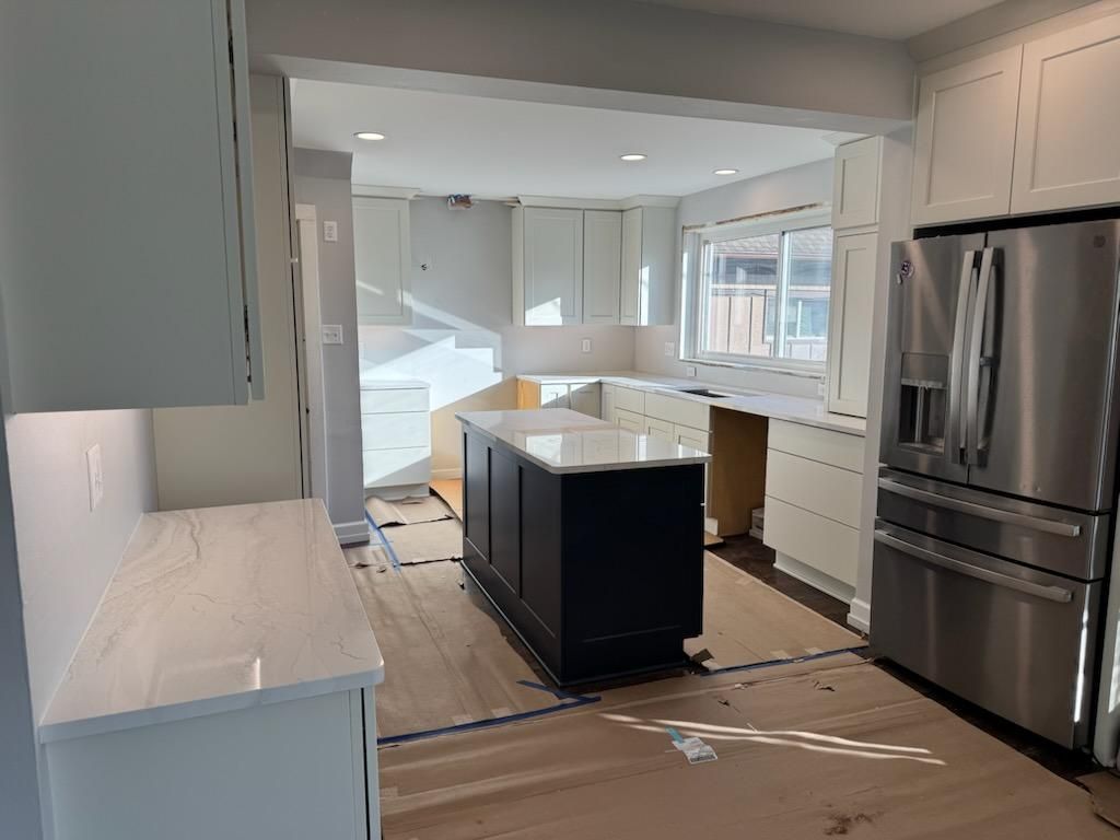 Bright kitchen under renovation with island, white cabinets, stainless fridge, and covered floor.