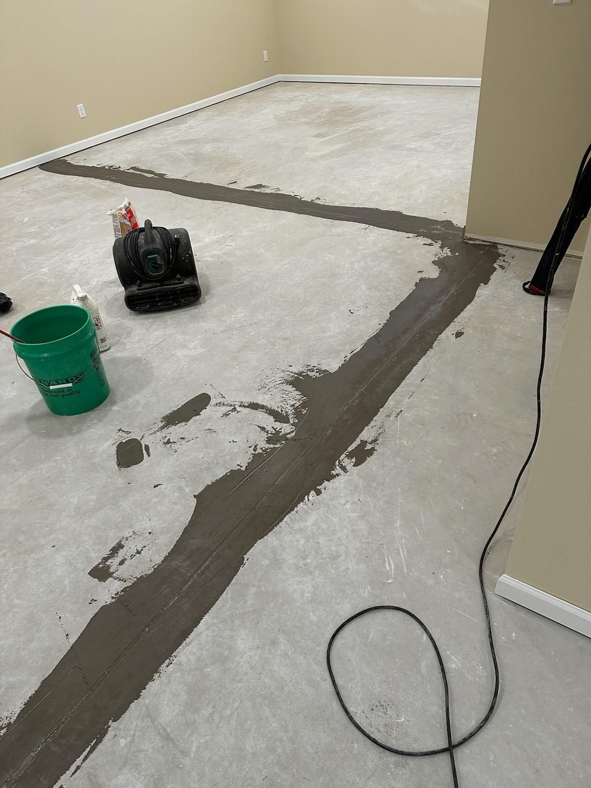 Partially painted floor with gray repair lines, green bucket, and paint supplies in an empty room