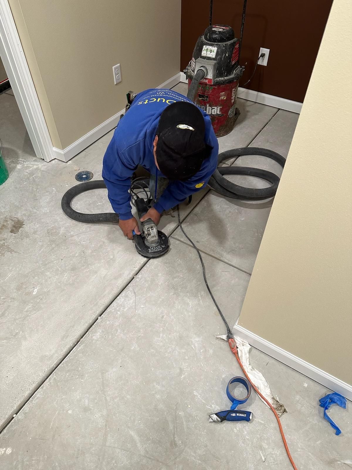 Worker in blue jacket using a floor tool on a concrete floor near cleaning equipment and a vacuum hose