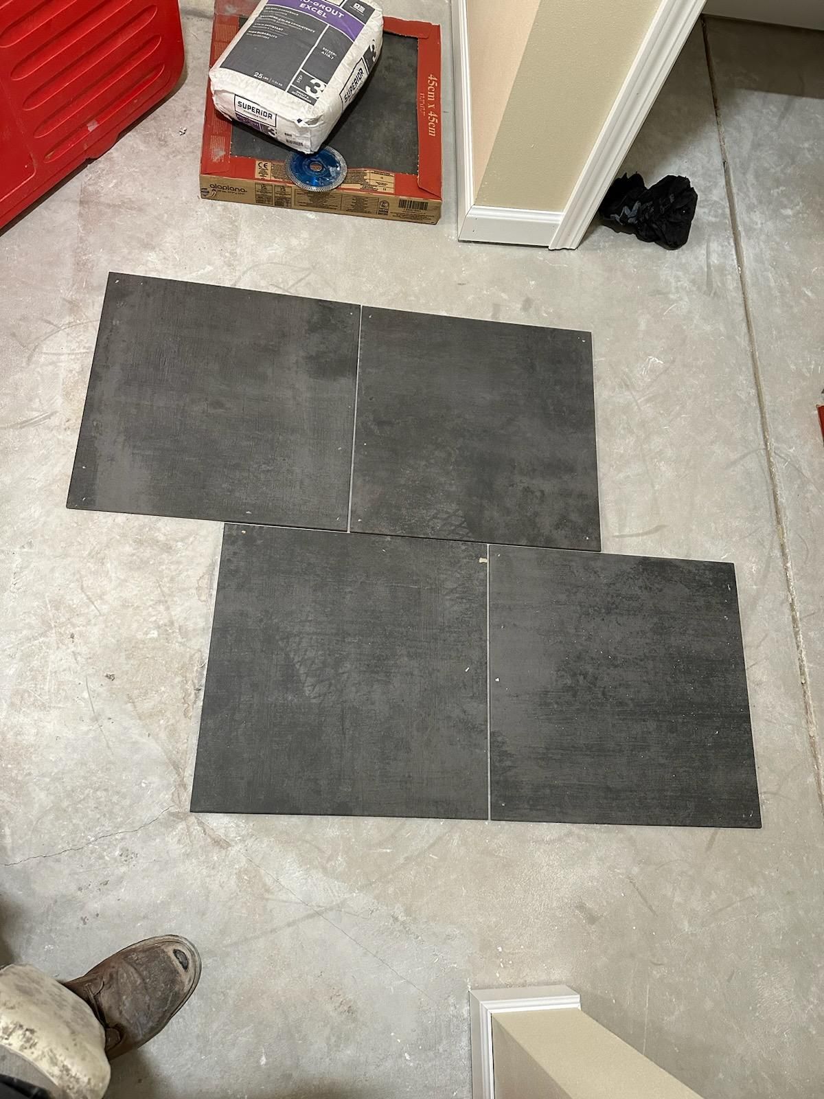 Four dark floor tiles laid out on a gray floor beside a wall, with a boot and small items nearby.
