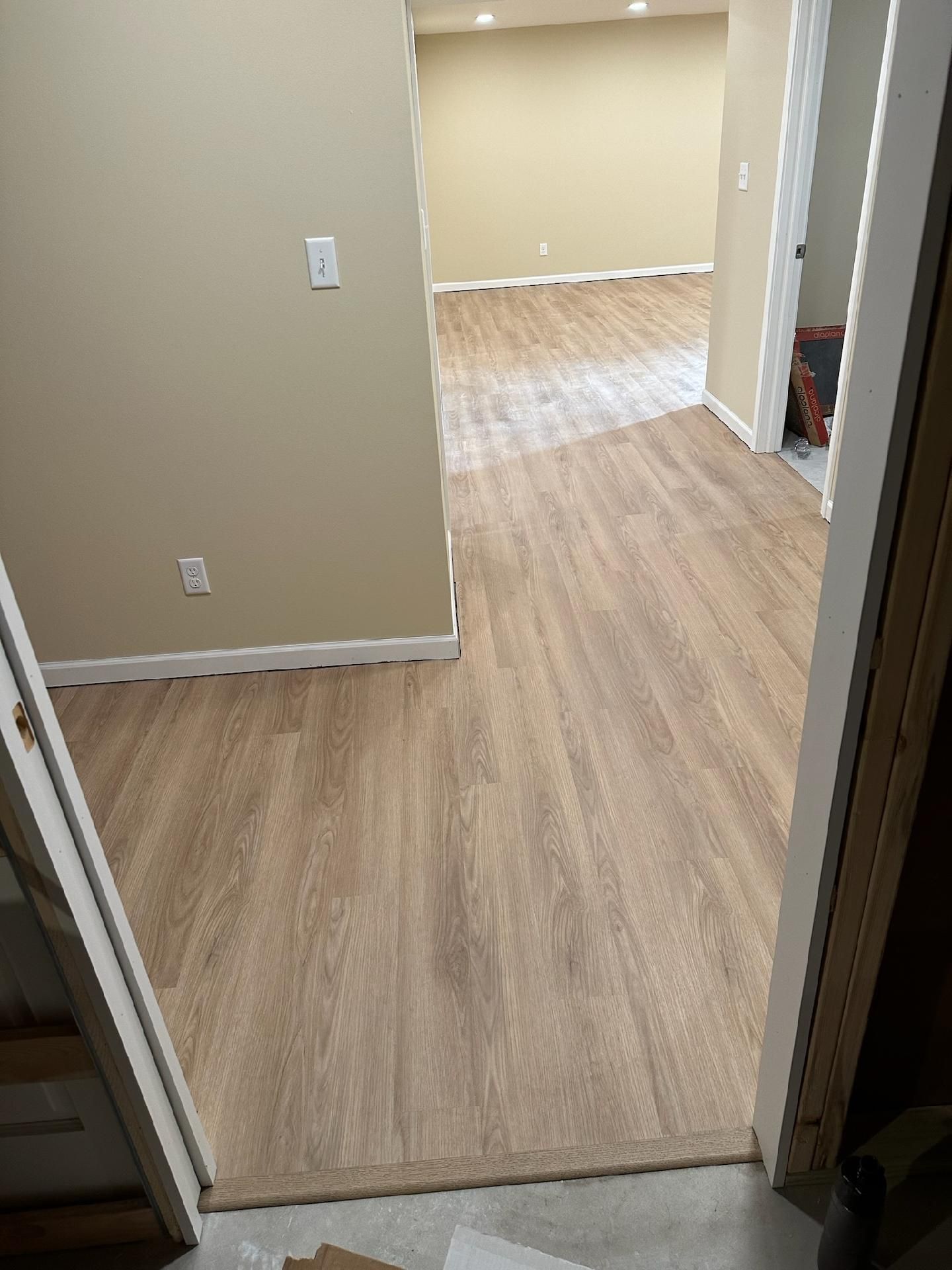 Empty hallway with beige walls and light wood flooring, leading into a room with matching floor.