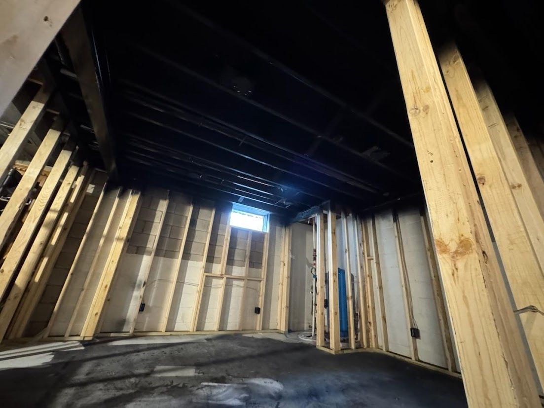 Empty unfinished room with exposed wooden studs, dark ceiling, concrete floor, and a small window opening.