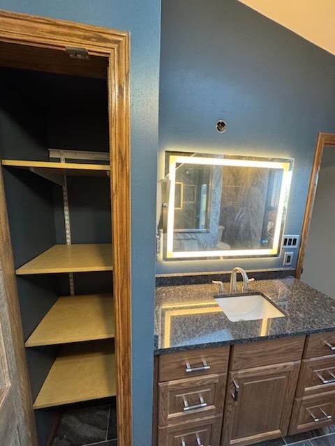 Bathroom vanity with lit mirror, granite countertop, sink, and open closet shelving beside it