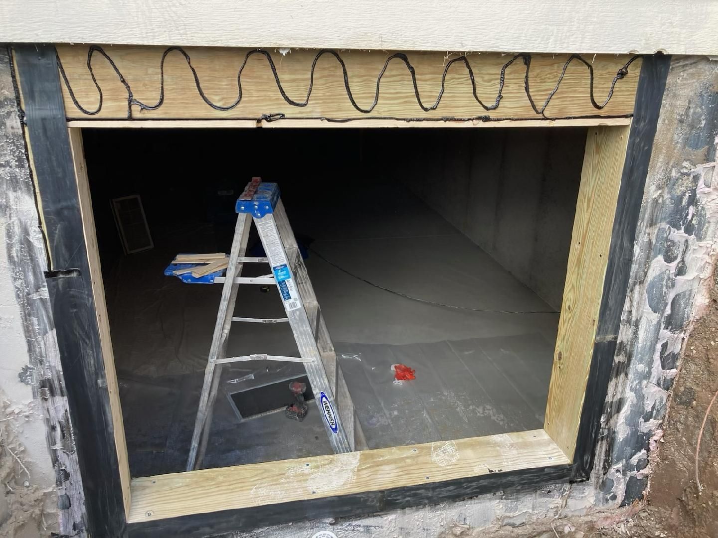 Ladder inside a dark basement opening beneath a garage, with exposed wiring and unfinished framing.
