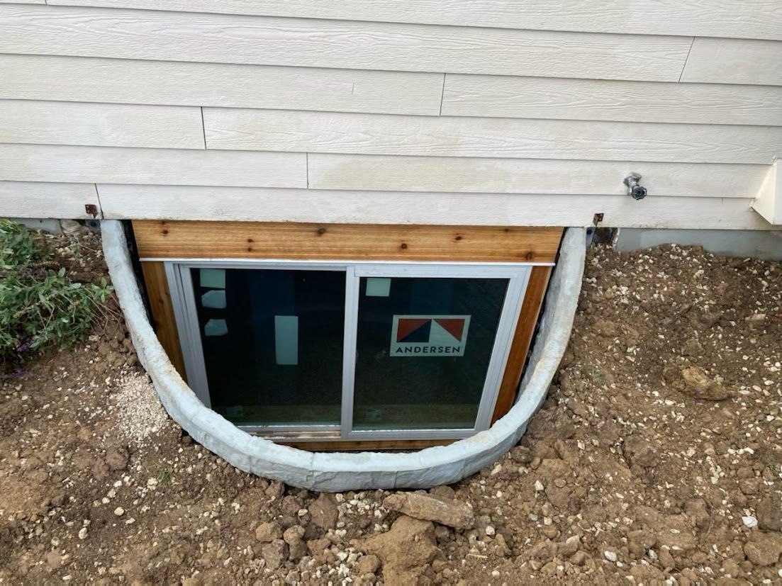 Basement window beneath house siding with a curved white pipe around it and exposed dirt in front