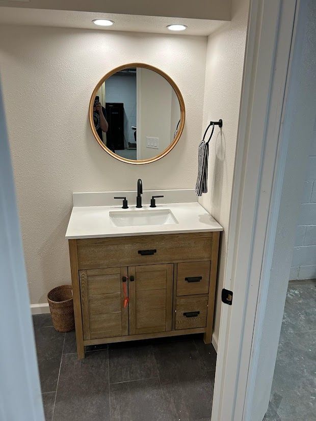 Small bathroom vanity with round mirror, white sink, wood cabinet, gray floor, and towel ring.