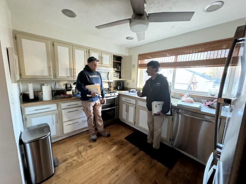 Two people standing in a bright kitchen, talking near the stove and sink.