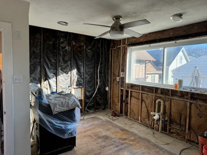 Partially gutted room with exposed studs, black plastic sheeting, and a large window letting in daylight