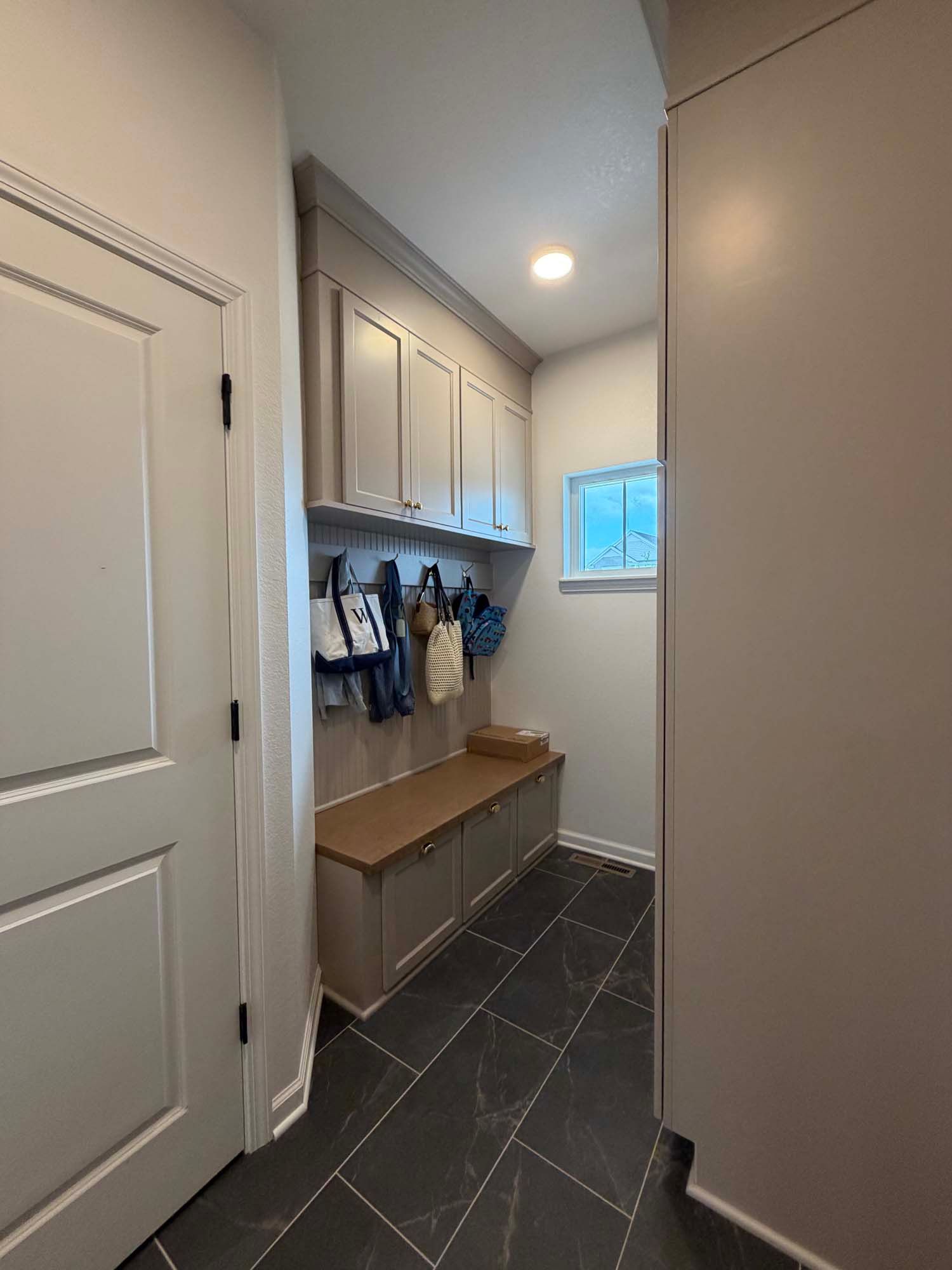 Narrow mudroom hallway with white cabinets, bench, hooks, gray tile floor, and a small window.