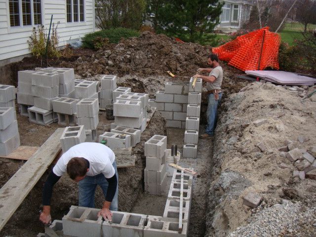 Workers building a cinder block foundation beside a house, with dirt piles and orange safety netting nearby