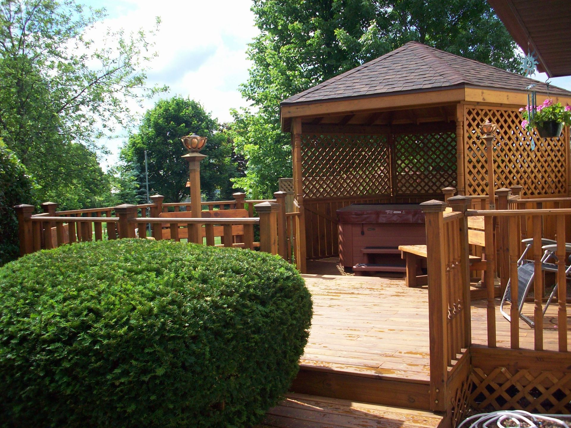 Wooden gazebo and deck in a leafy backyard, with a trimmed bush in the foreground.