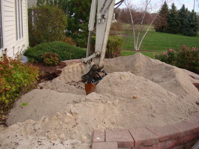 Excavator digging a large sand pile beside a house in a landscaped yard