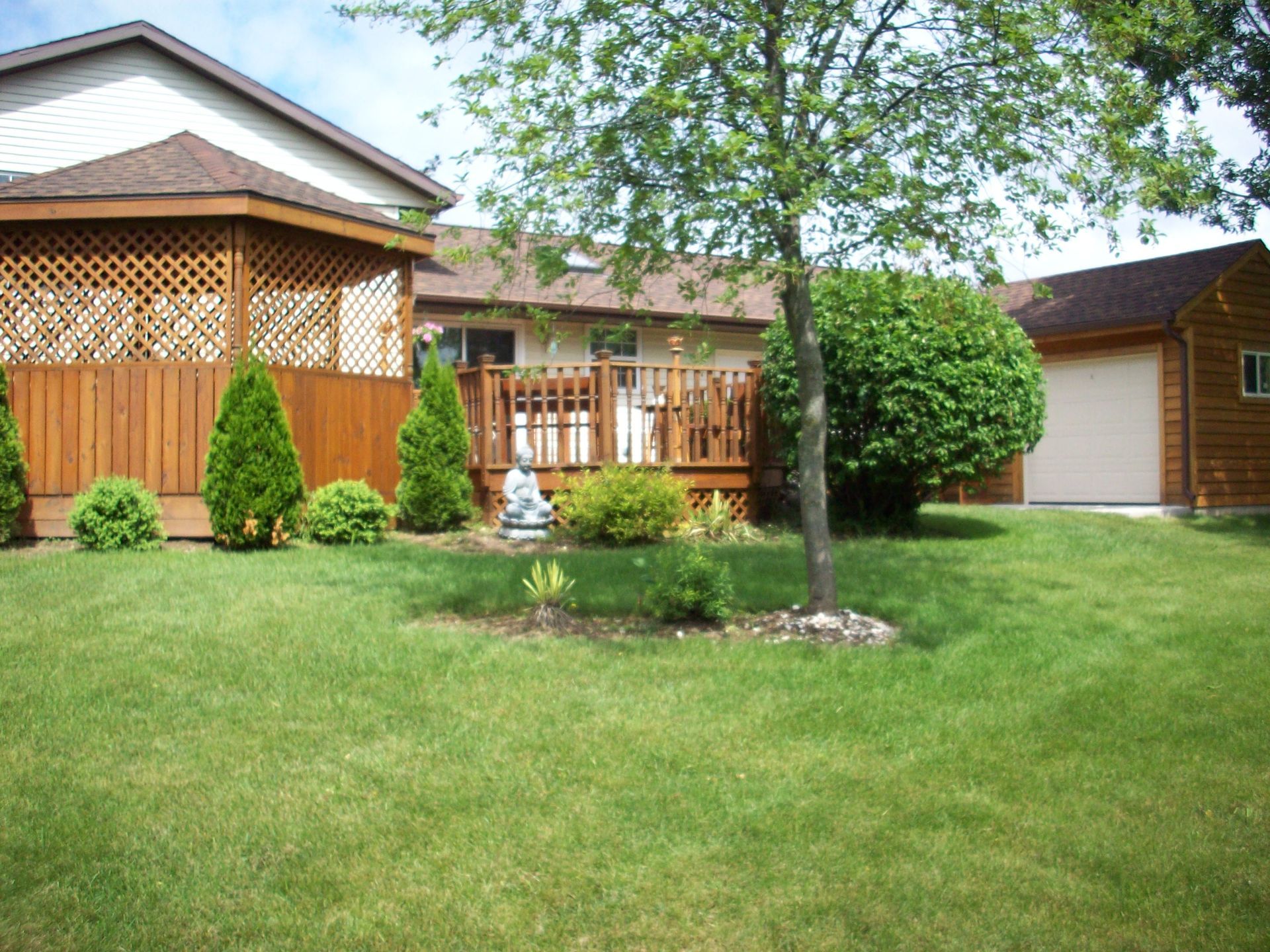 Backyard with green lawn, wooden house, deck, trees, and shrubs under a bright sky