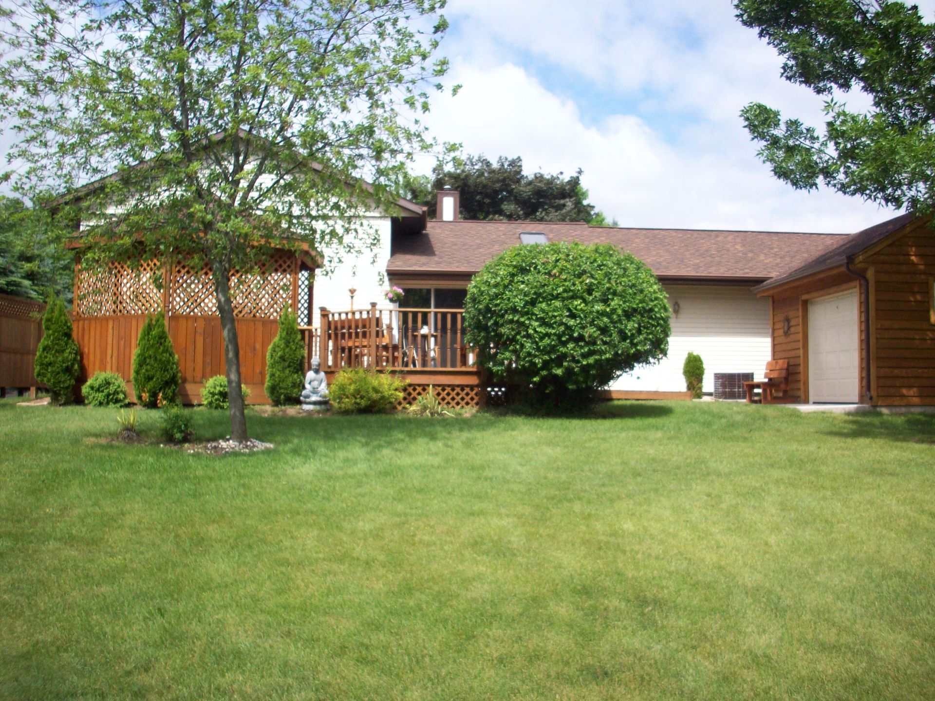 Suburban house with a large green lawn, trees, and a wooden deck under a partly cloudy sky