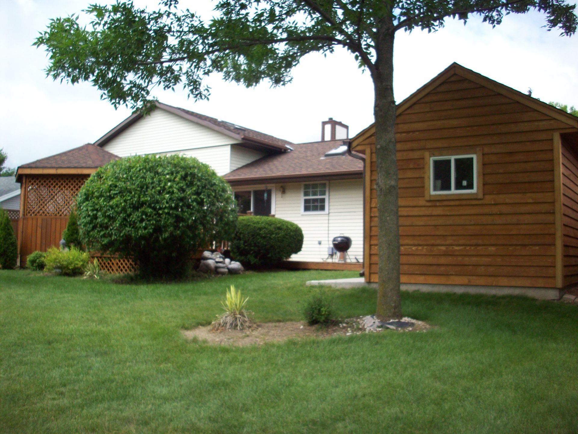 Suburban backyard with green lawn, trees, bushes, and two small wooden sheds near a white house