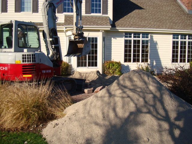 Excavator beside a house, digging near a large mound of dirt in a front yard.