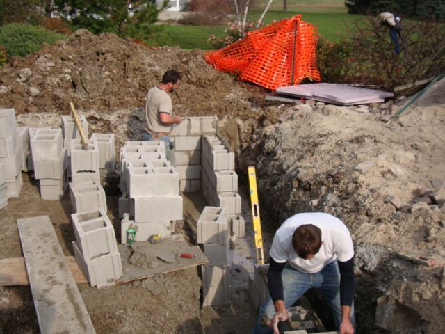 Workers laying cinder blocks at a muddy construction site with orange safety netting nearby