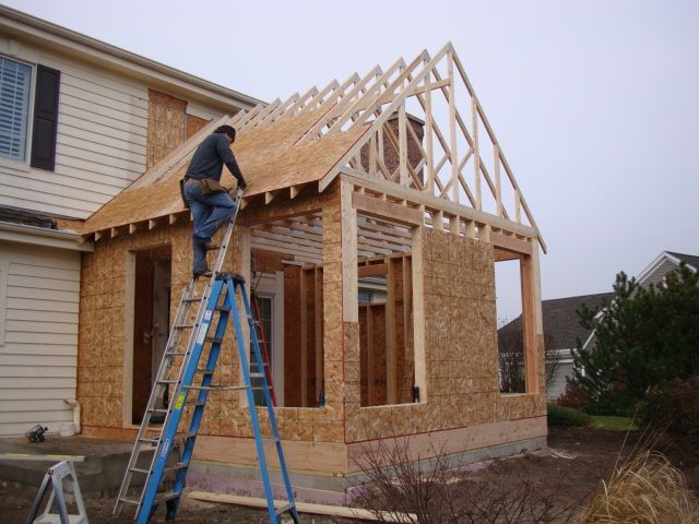 Worker framing a house addition on a blue ladder, installing roof trusses on a partly built brick exterior.