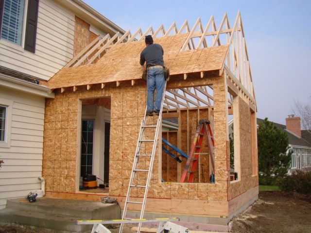 Worker building a house addition with exposed wooden framing and a ladder on site