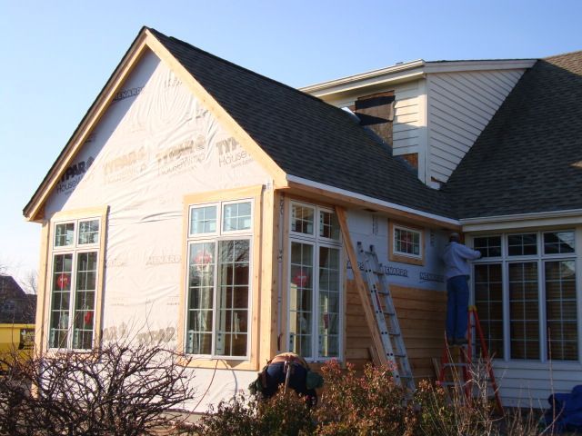 Workers installing siding on a house addition, with a ladder and one person on the roof.