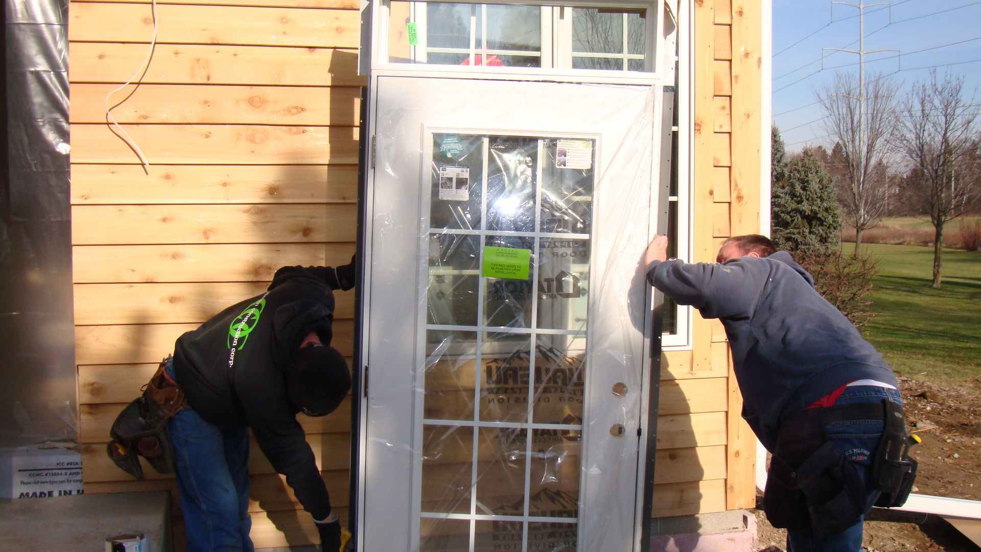 Two people work on the exterior of a small wooden building beside a glass door.