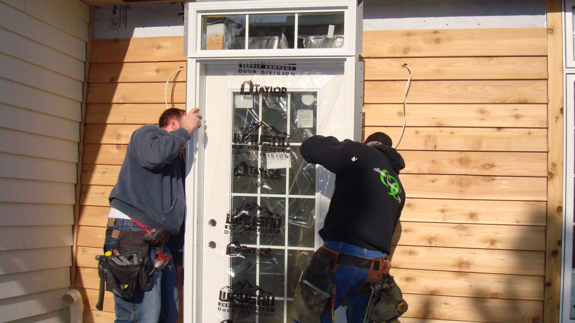 Two workers installing trim around a white door on a house exterior.