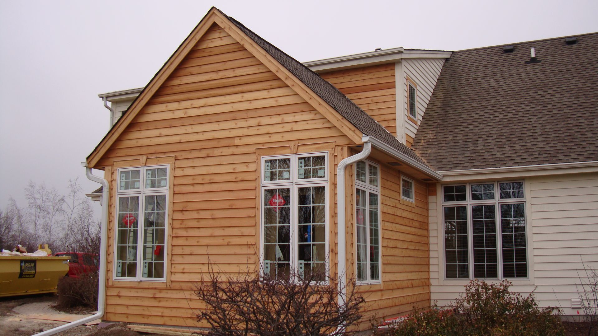 Front view of a tan wooden house addition with tall windows and a gray shingled roof on a cloudy day