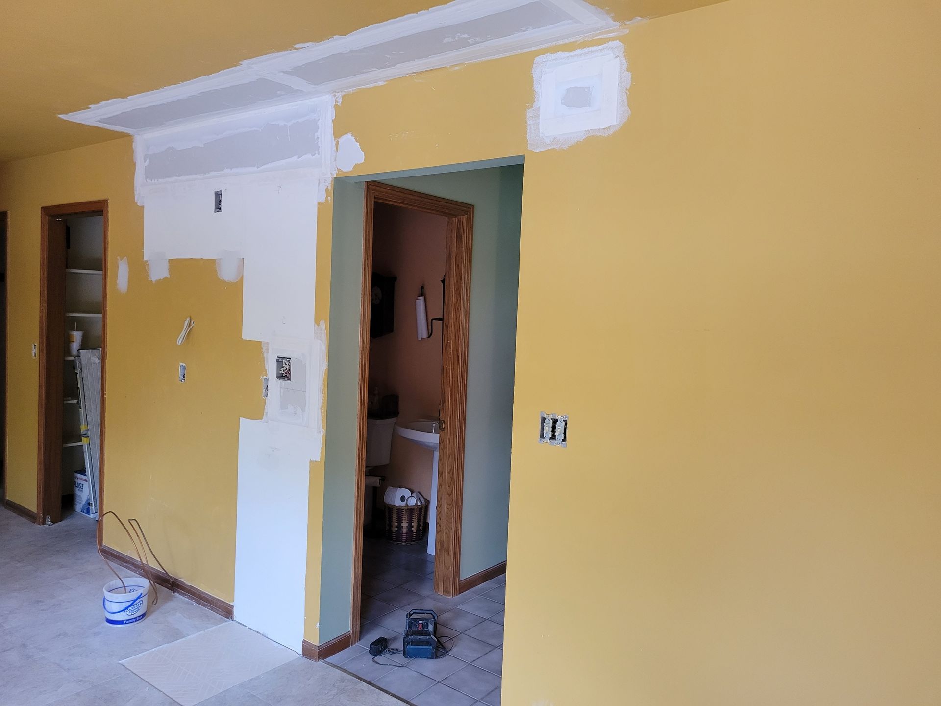 Yellow room under renovation with white drywall patches, open doorway, and paint bucket on the floor