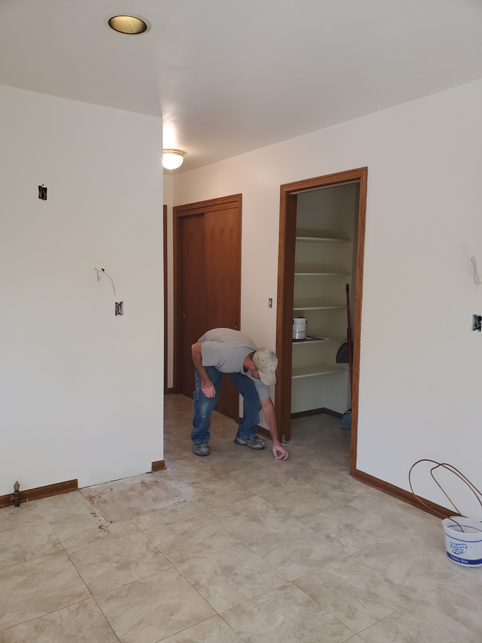 Person bending over in a nearly empty room with white walls, brown doors, and a tiled floor.