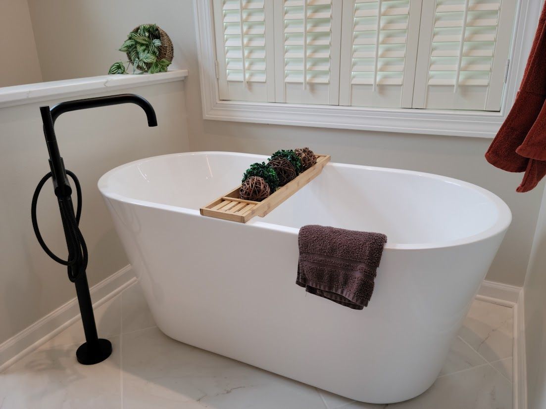 Modern white freestanding bathtub beside a black floor faucet, with a bath tray and towel in a bright bathroom