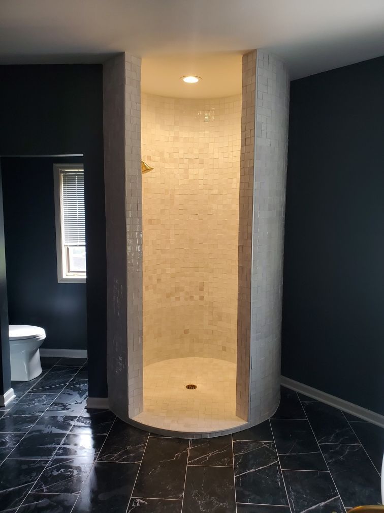 Small tiled shower stall in a dark bathroom with black floor tiles and a window nearby